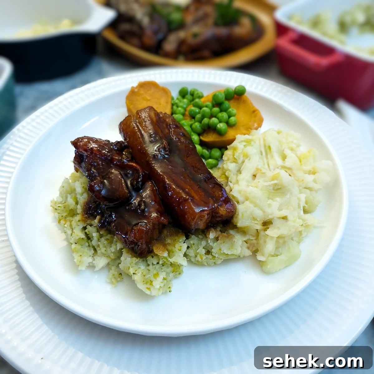 Two glazed pork belly slices on a pile of vegetable mash on a white plate with sweet potatoes and peas.