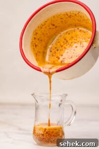 Close-up of homemade hot honey being poured from a small saucepan into a glass pitcher, showing its golden color speckled with red pepper flakes.