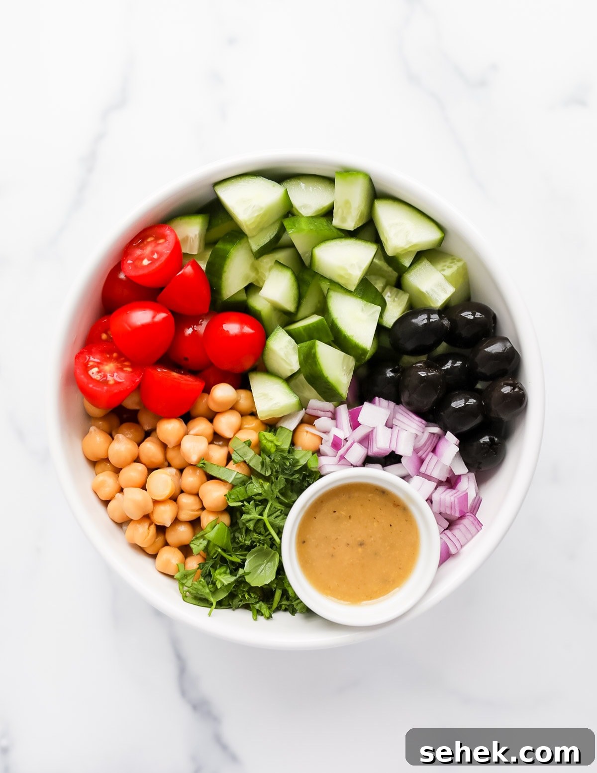 Classic Greek Cucumber Salad with Feta and Olives 5 A bowl of unmixed ingredients with sectioned off ingredients including: tomatoes, cucumber, olives, red onion, dressing, herbs, chickpeas, and tomatoes.