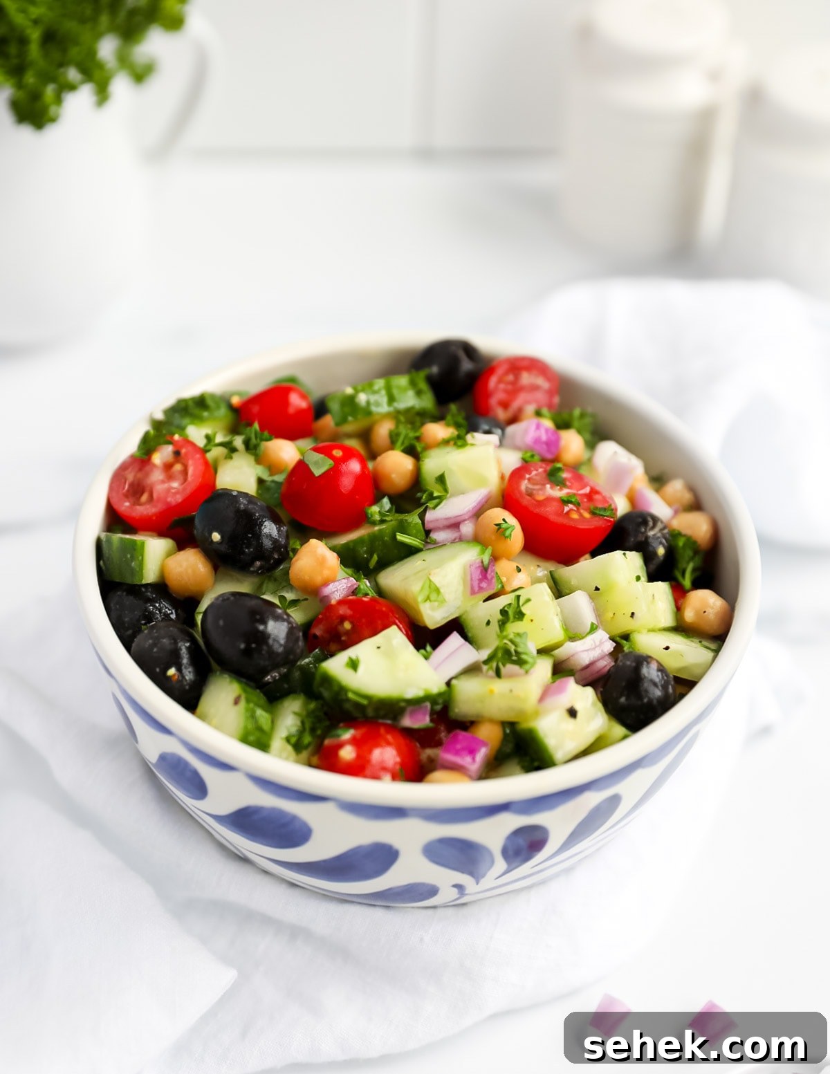 Classic Greek Cucumber Salad with Feta and Olives 7 A blue and white bowl filled with a Greek salad including olives, tomatoes, chickpeas, cucumbers, and onion, covered in fresh herbs