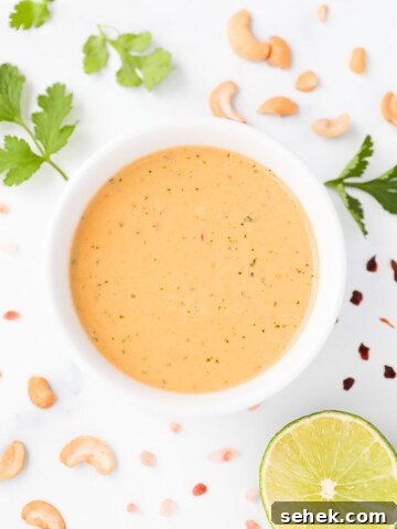 A creamy orange salad dressing in a white bowl. There are supporting ingredients around the bowl including: cilantro, cashews, red pepper flakes, a sliced lime, and salt