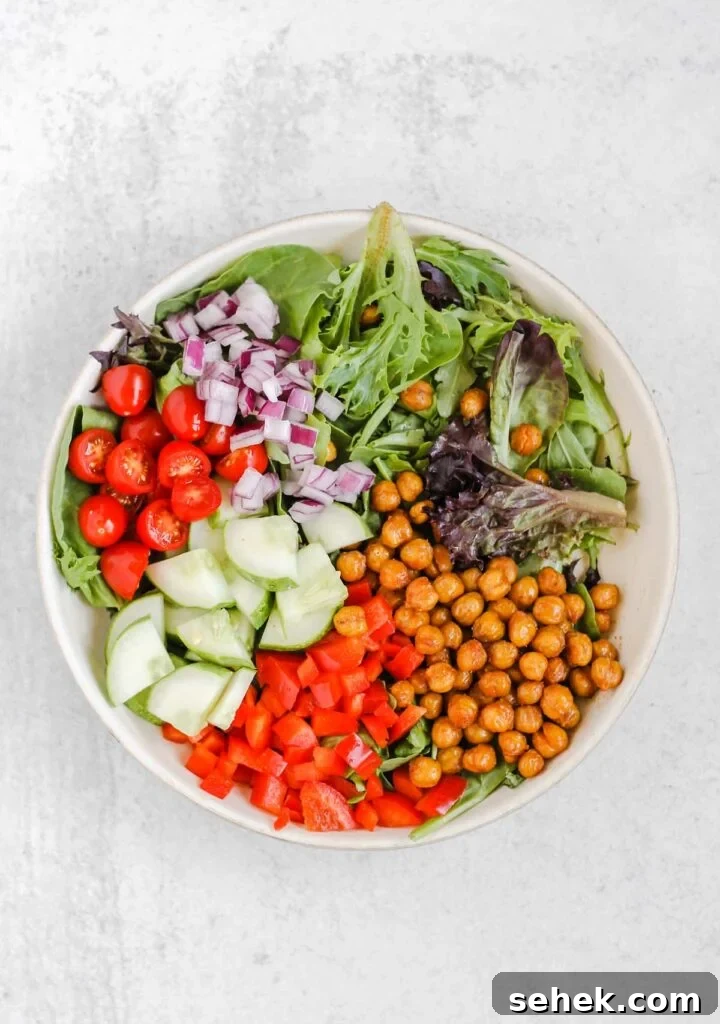 Mixed greens, roasted chickpeas, peppers, cucumbers, tomatoes, and onions in a salad bowl.
