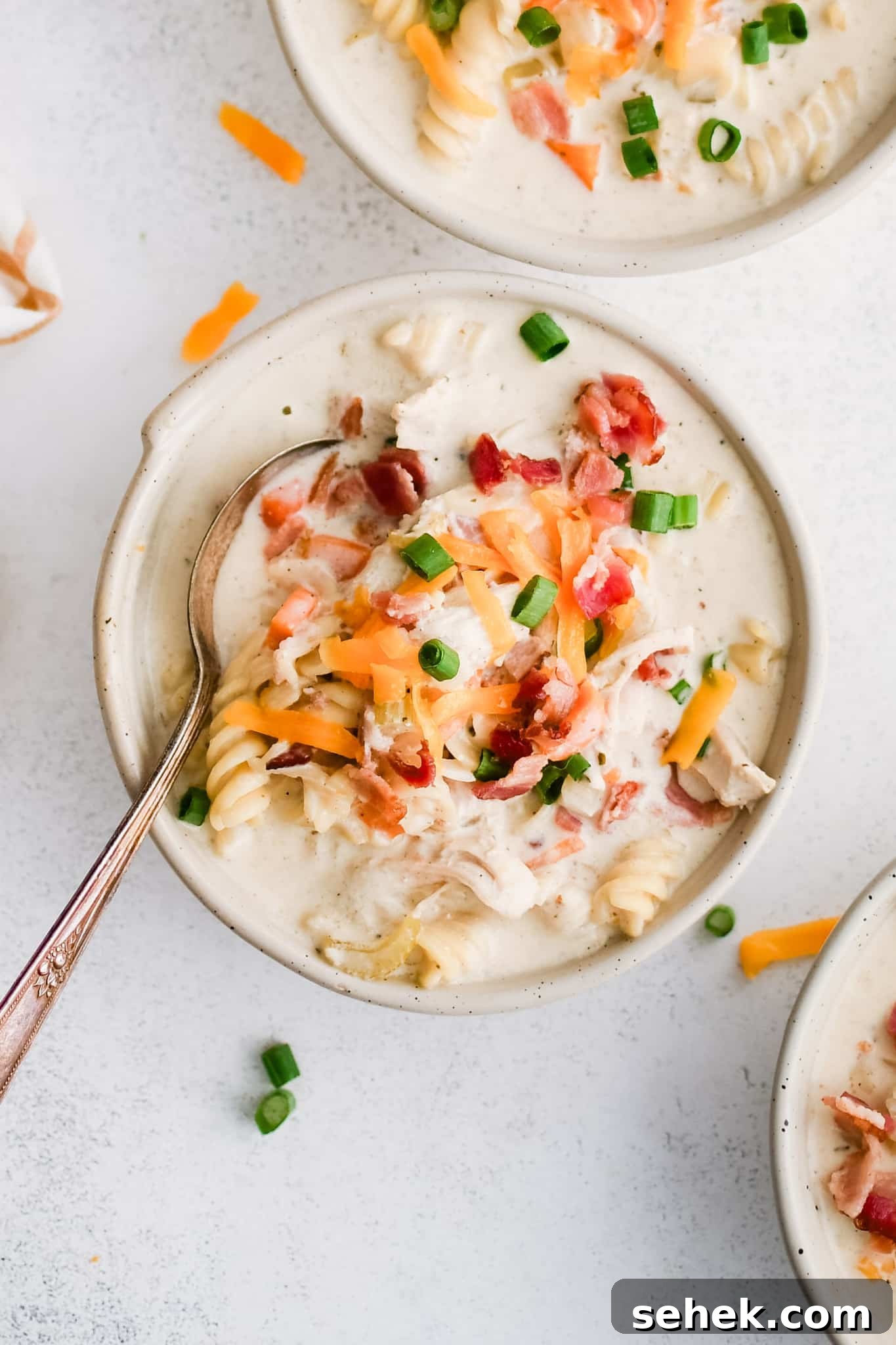 Three white soup bowls filled with super creamy crack chicken soup filled with rotini pasta, shredded chicken, bacon, and topped with shredded cheddar cheese, chopped green onions, and even more crispy cooked bacon bits.