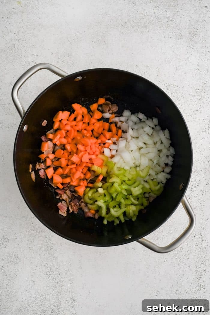 Large soup pot filled with crispy cooked bacon bits covered by diced carrots, celery, and onions.