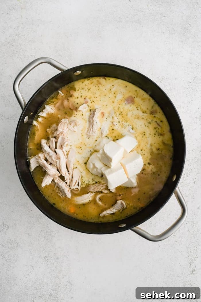 Shredded chicken, cubed cream cheese, and heavy cream added to a large soup pot filled with simmering pasta, bacon. and veggies in broth.