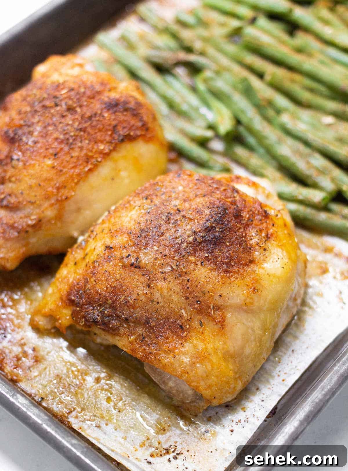 Close up of baked bone-in skin-on chicken thigh coated in seasoning with green beans in the background.
