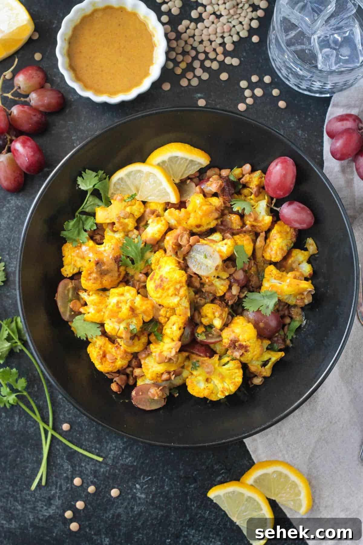 Roasted Cauliflower Salad - cauliflower tossed with turmeric & curry powder, roasted until tender and golden, and then tossed with protein-packed lentils and sweet, juicy grapes. It's flavor and texture dynamite! A light, yet filling, meatless meal or side dish. Curry roasted cauliflower salad in a black serving bowl garnished with two lemon slices.