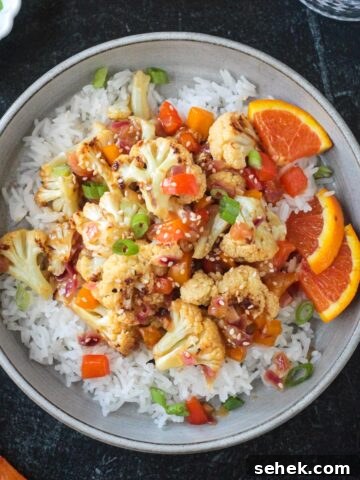 Vegan orange cauliflower in a sticky sauce with diced peppers over rice in a gray flat bowl.