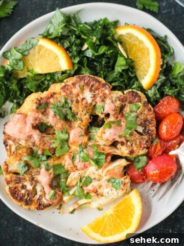 One roasted cauliflower steak garnished with spicy yogurt sauce and chopped parsley on a plate with kale salad and tomatoes.