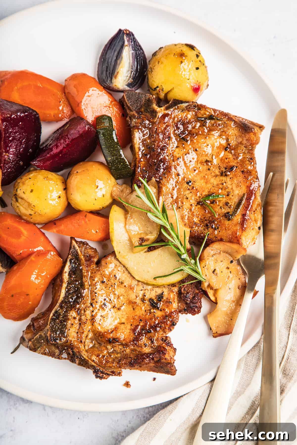 A plate of baked pork chops with apples and winter vegetables, garnished with a sprig of rosemary.