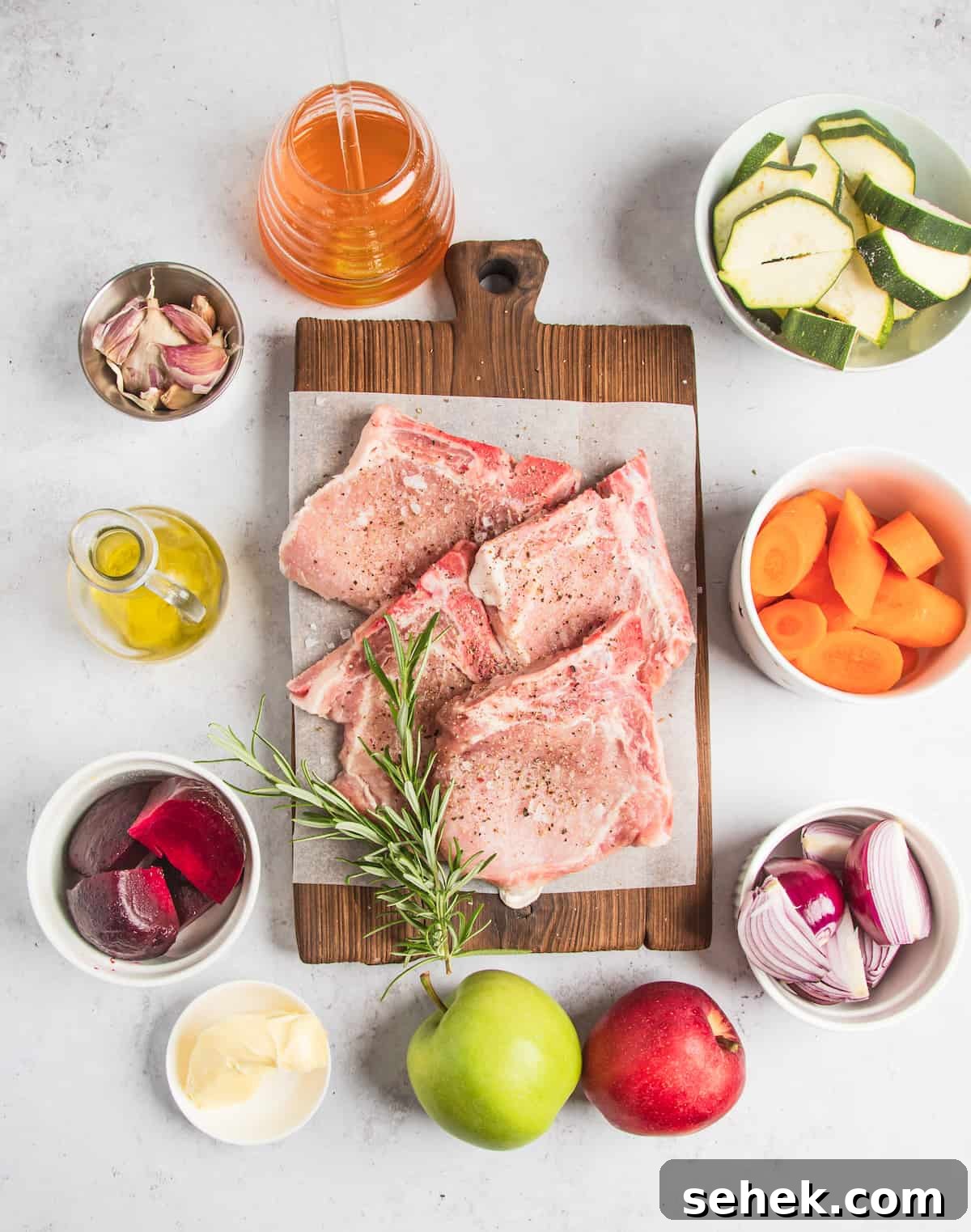 Clockwise from top: Sliced zucchini, chopped carrots, chopped onions, two whole apples, butter, beetroot, oil, garlic, and apple cider.  In the center: Four seasoned pork chops and a sprig of fresh rosemary.
