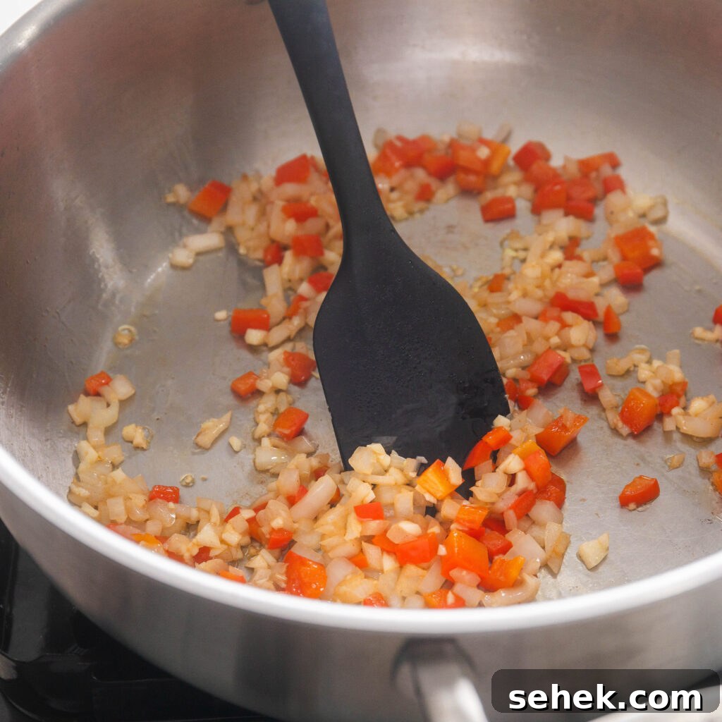 Sautéing diced onion, red bell pepper, and garlic in a stainless steel sauce pan with a black silicone spatula.