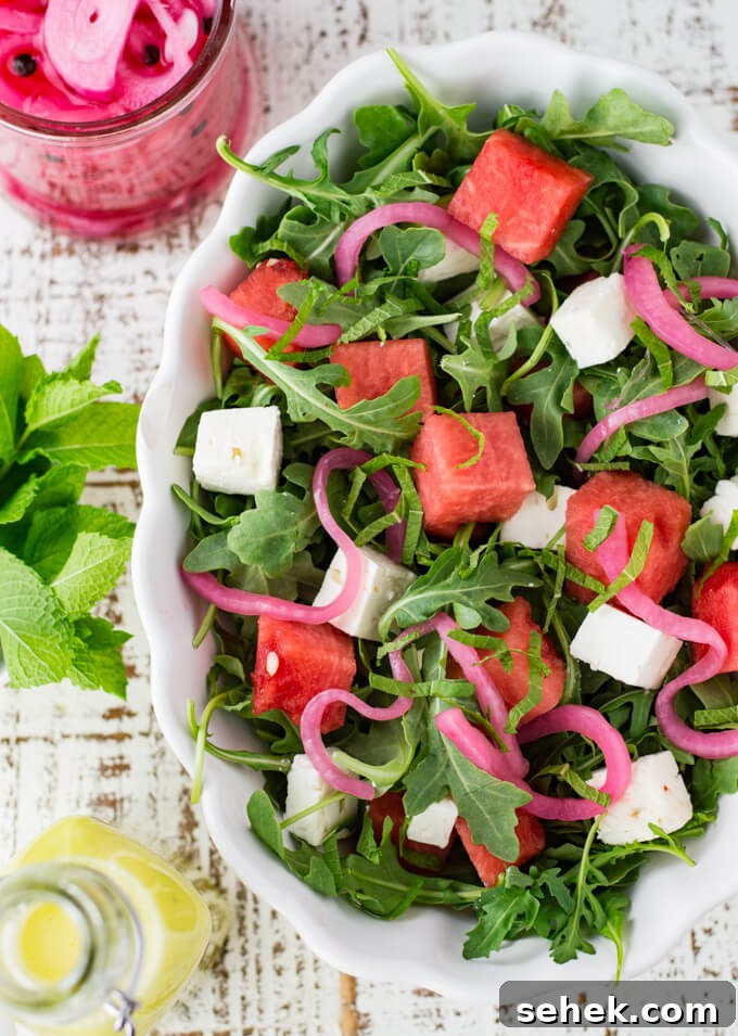 Watermelon Arugula Salad with Feta & Mint 2 A white serving bowl with watermelon arugula salad. Feta cheese and pickled onion are in the salad as well. It's drizzled with poppyseed dressing. Mint, a bottle of dressing and pickled onions sit next to the bowl.