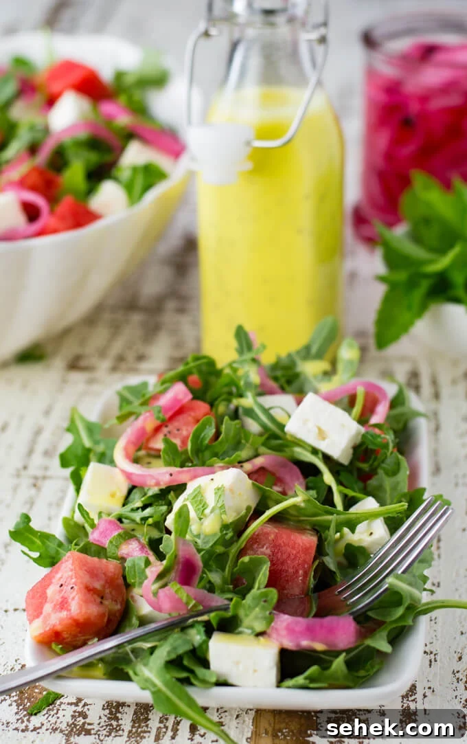 Watermelon Arugula Salad with Feta & Mint 7 A white plate with watermelon arugula salad. Feta cheese and pickled onion are in the salad as well. It's drizzled with poppyseed dressing. Mint, a bottle of dressing and pickled onions sit next to the bowl.