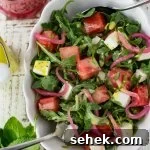 A white serving bowl with watermelon arugula salad. Feta cheese and pickled onion are in the salad as well. It's drizzled with poppyseed dressing. Mint, a bottle of dressing and pickled onions sit next to the bowl.