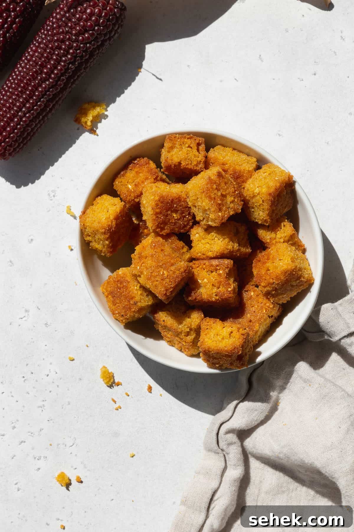 Crispy Cornbread Croutons for Salads and Soups 2 An overhead shot of a small bowl of cornbread croutons with a napkin and a stalk of heirloom corn.