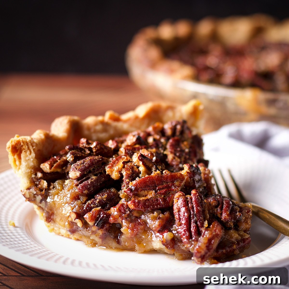 A slice of pecan pie on a white plate with a gold fork resting next to it.
