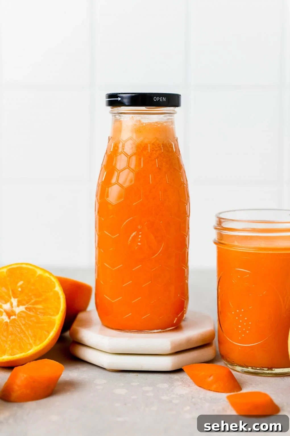 Carrot, Apple and Orange Juice with Ginger 2 Straight on shot of a textured glass jar filled to the brim with bright orange carrot, apple, and orange juice with ginger. Next to some cut oranges, and carrot pieces, on a light colored background.