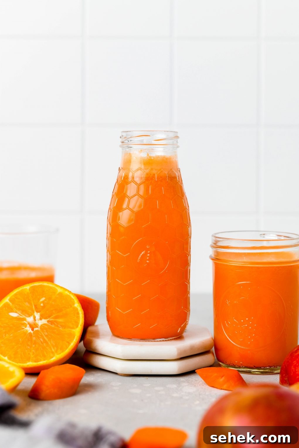 Carrot, Apple and Orange Juice with Ginger 7 Straight on shot of a textured glass jar filled to the brim with bright orange carrot, apple, and orange juice with ginger. Next to some cut oranges, and carrot pieces, on a light colored background.