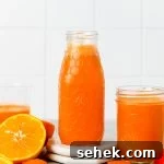 A glass bottle and a jar filled with bright orange juice sit on a table, surrounded by fresh orange halves and carrot pieces. The background is a white tiled wall.