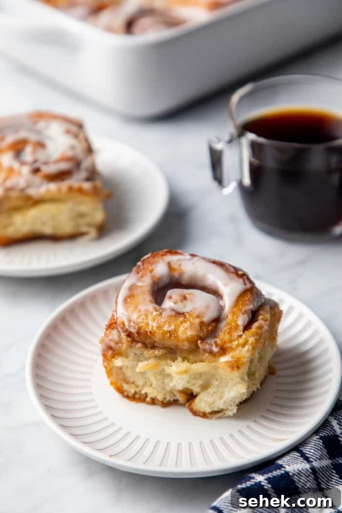 TikTok Cinnamon Rolls Made with Heavy Cream 3 Frosted cinnamon roll with heavy cream on a white plate. A second cinnamon roll and a cup of coffee are in the background.