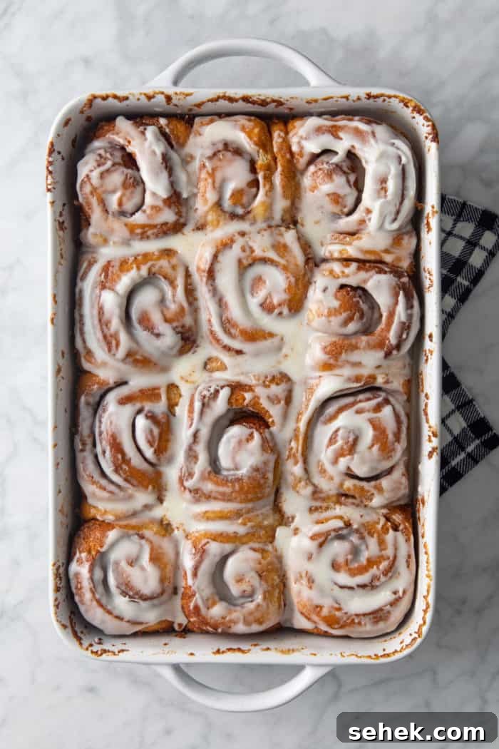 TikTok Cinnamon Rolls Made with Heavy Cream 8 Frosted cinnamon rolls with heavy cream in a white baking dish, resting on a tea towel to cool.