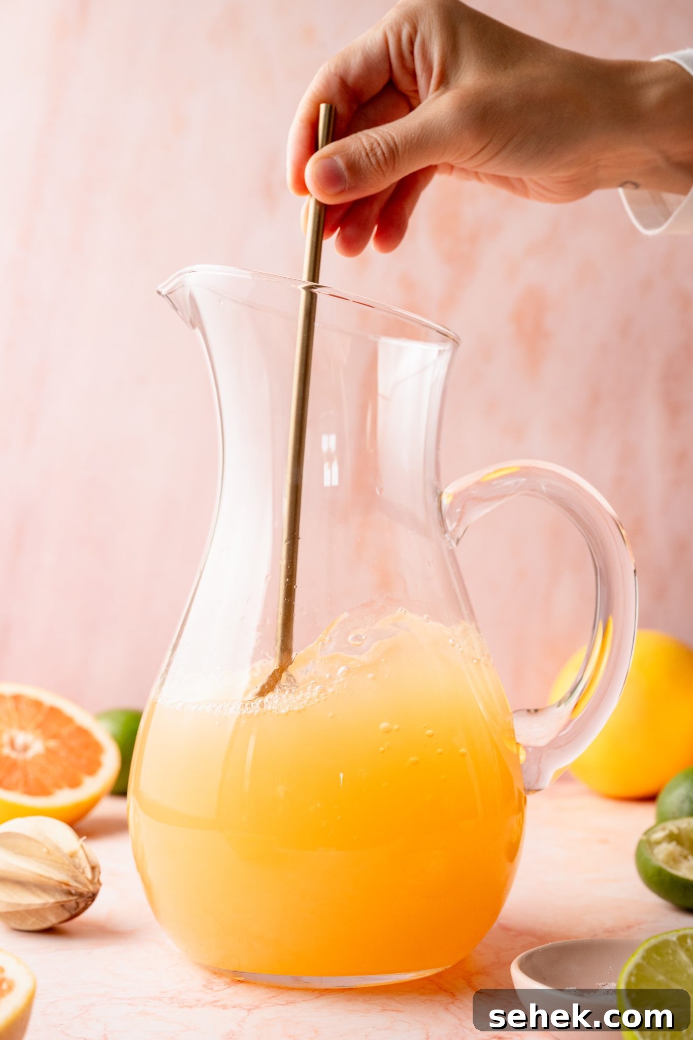 Paloma Pitcher: Grapefruit Tequila Cocktail for Parties 10 A hand stirs a large clear pitcher of freshly mixed Paloma cocktail with a golden stirring stick. Cut citrus and cocktail ingredients are scattered around the pitcher on a pink background.