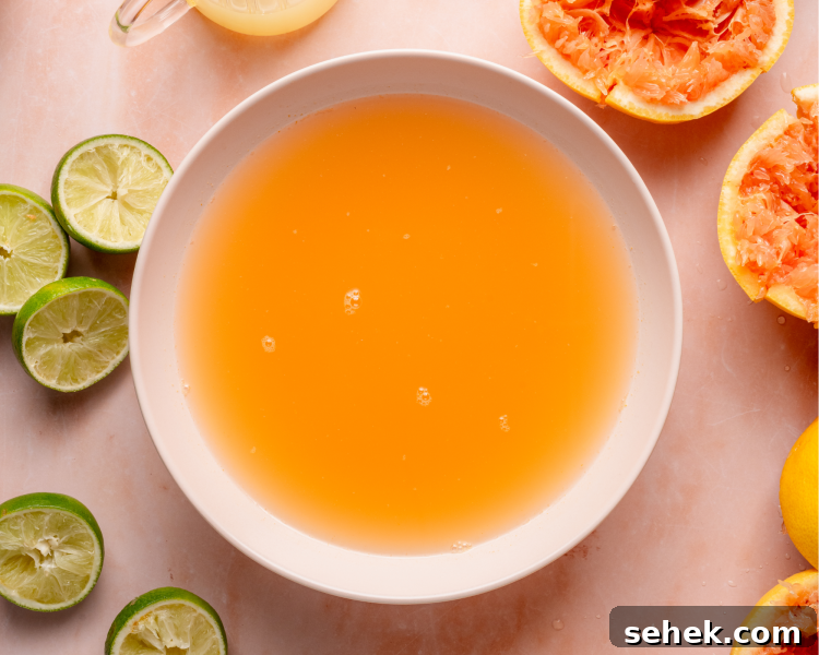 Overhead shot of a large bowl filled with fresh grapefruit-lime juice. Squeezed citrus halves, lime slices, and a small pitcher of juice surround the bowl on a pink marble surface.