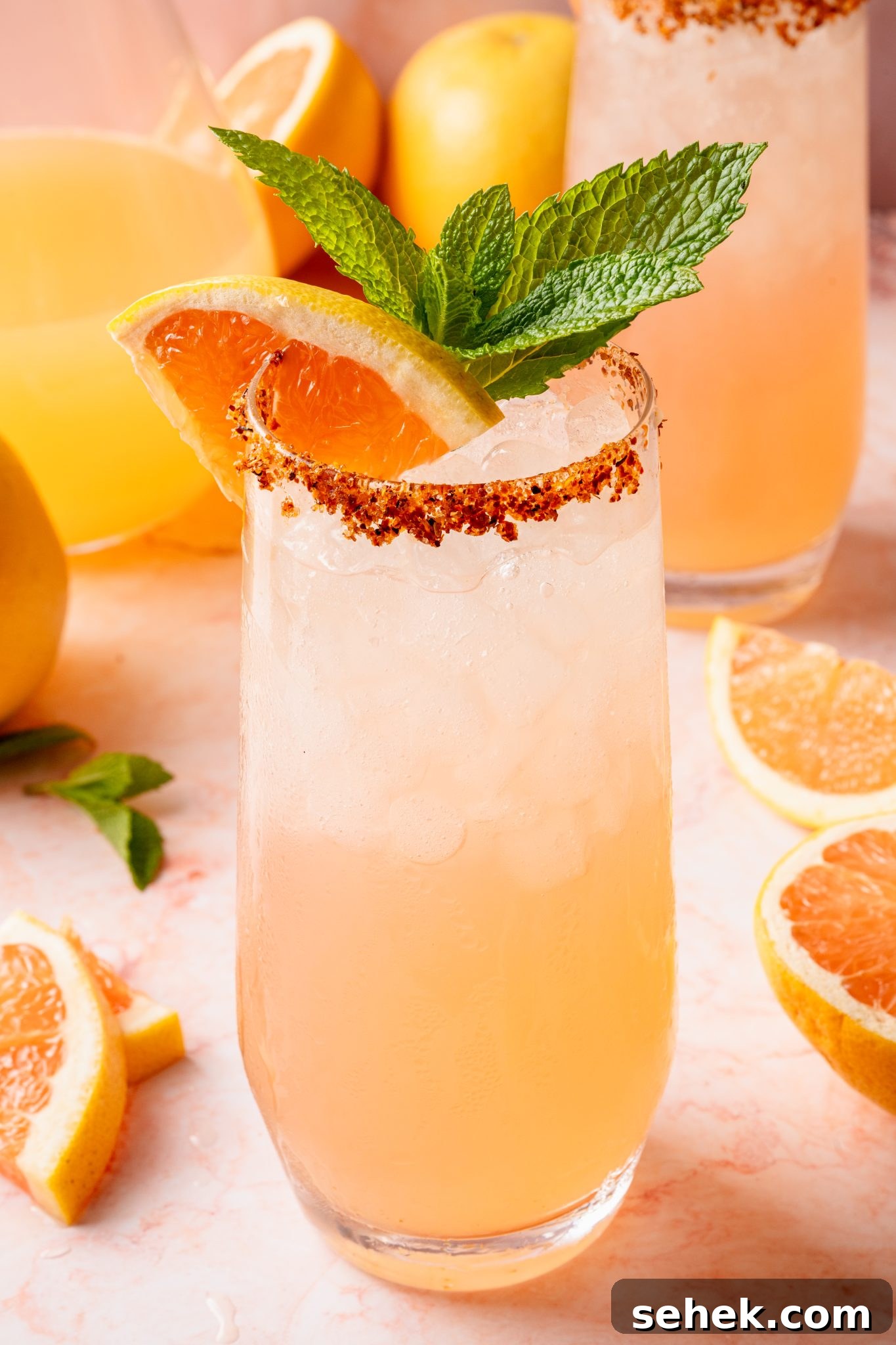 Paloma Pitcher: Grapefruit Tequila Cocktail for Parties 14 A tall glass of pink Paloma cocktail filled with crushed ice, garnished with a grapefruit wedge, lemon slice, and fresh mint, with a chili-lime salt rim. A pitcher of Paloma and another garnished glass are visible in the background alongside citrus fruits and mint on a pink marble surface.