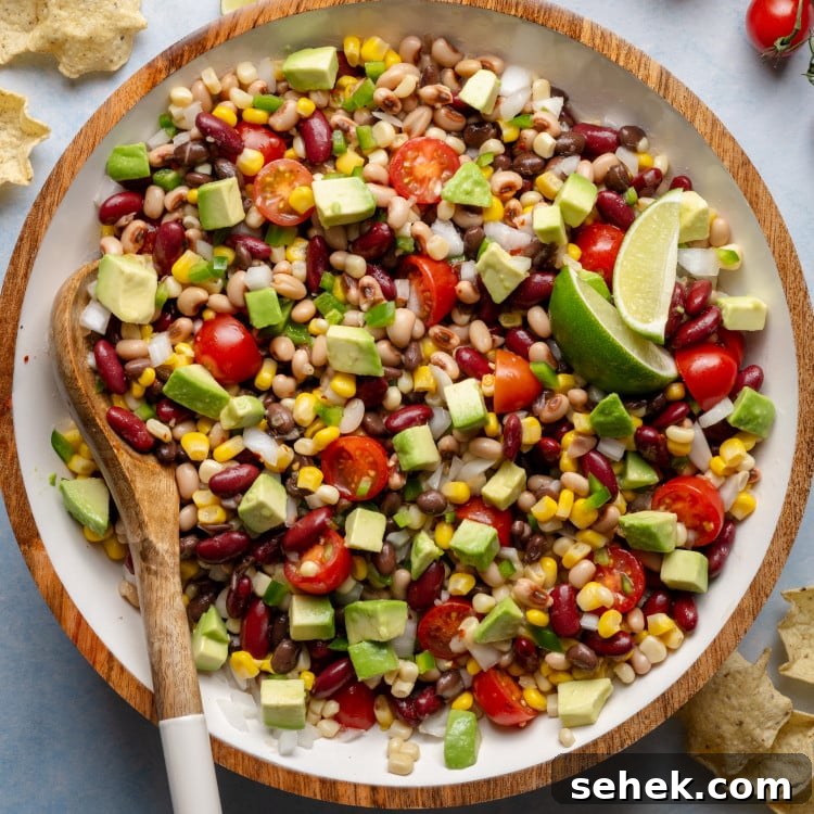 Mixed cowboy caviar in a large bowl with a wooden spoon, showing vibrant beans, corn, tomatoes, jalapeños, and diced avocado.