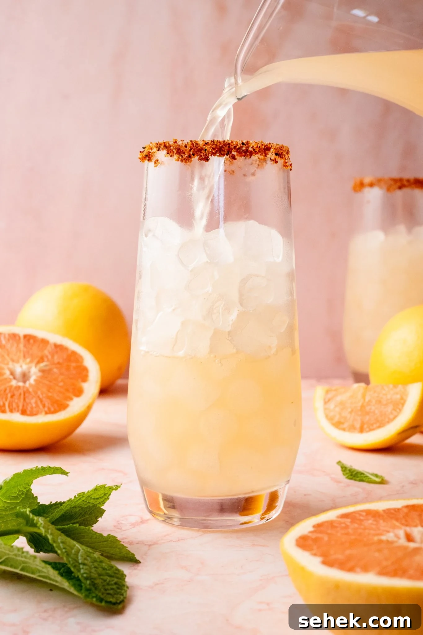 Paloma Pitcher: Grapefruit Tequila Cocktail for Parties 8 A glass with a chili-lime rim being filled with grapefruit Paloma cocktail over ice from a clear pitcher. The background includes sliced grapefruit, lemons, and mint leaves on a pink marble surface.