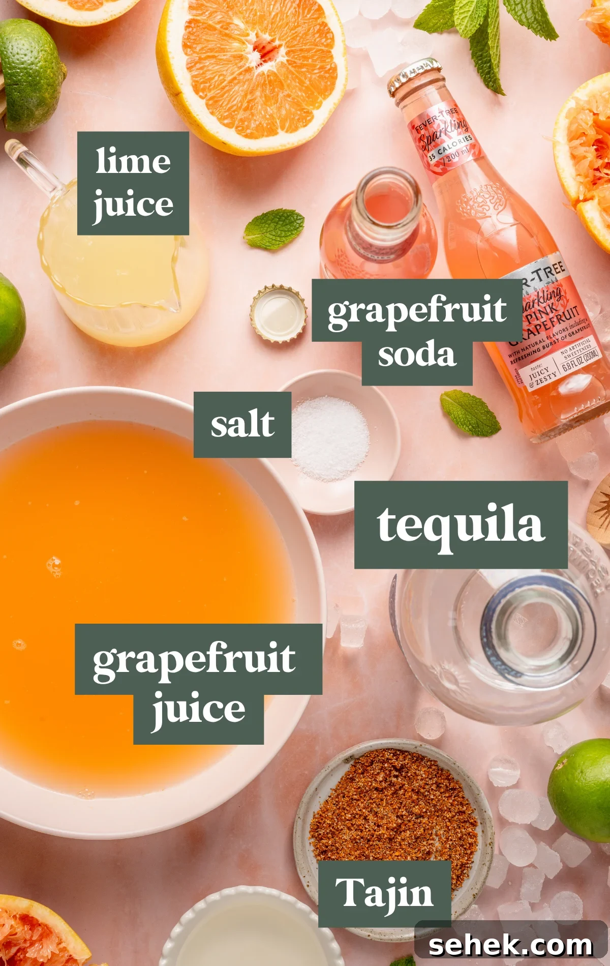 Paloma Pitcher: Grapefruit Tequila Cocktail for Parties 9 Overhead shot of Paloma cocktail ingredients including halved grapefruits, limes, grapefruit soda, lime juice, chili-lime salt, a bowl of orange-hued juice, and fresh mint on a pink surface with scattered ice.